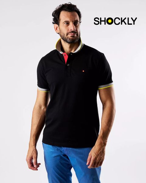 Ein Mann in einem schwarzen Poloshirt mit gelb-weißen Akzenten und einer blauen Hose steht vor einem schlichten weißen Hintergrund. Oben rechts steht in schwarzer Schrift das Wort „SHOCKLY“.