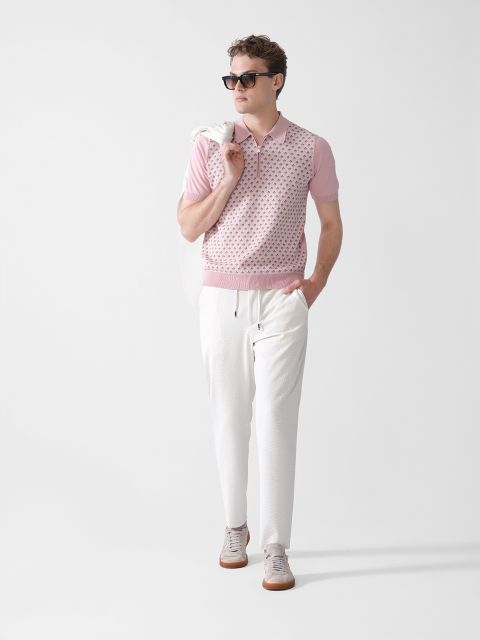 Ein Mann mit Sonnenbrille, einem rosa gemusterten Kurzarm-Poloshirt, weißen Hosen und hellen Turnschuhen steht mit einer Hand in der Tasche und einem weißen Pullover über der Schulter vor einem schlichten weißen Hintergrund.