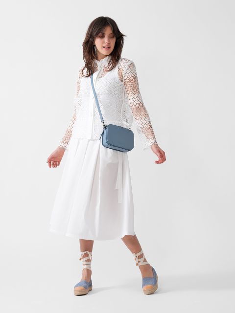 Eine Frau, die eine weiße Langarmbluse aus Spitze, einen weißen Midirock, blaue Espadrilles mit Knöchelbändern und eine hellblaue Umhängetasche trägt, steht vor einem schlichten weißen Hintergrund.