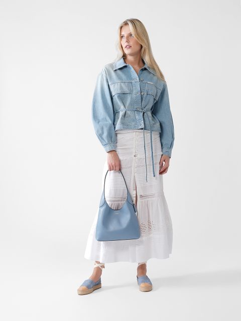 Vor einem weißen Hintergrund steht eine Frau, die eine helle Jeansjacke, einen langen weißen Rock mit Ösendetails und hellblaue Slipper trägt und eine passende hellblaue Handtasche in der Hand hält.