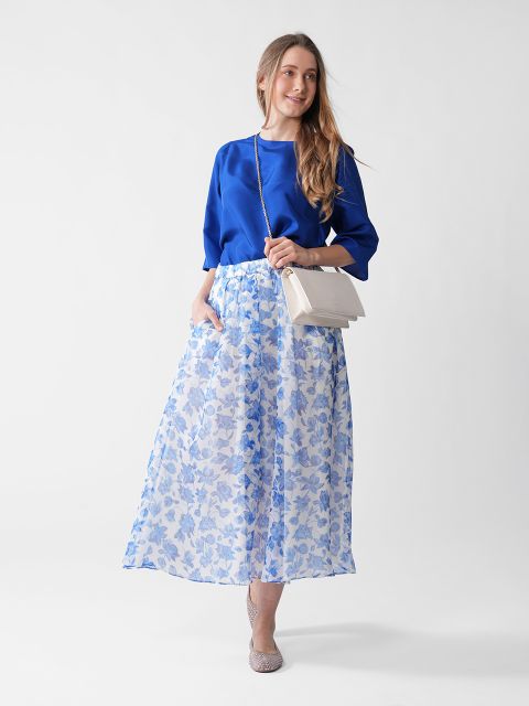 Eine Frau steht vor einem weißen Hintergrund. Sie trägt eine leuchtend blaue Bluse und einen langen, fließenden, weiß-blauen Rock mit Blumenmuster. Sie trägt eine helle Umhängetasche und gemusterte Schuhe. Ihr Haar ist locker frisiert.
