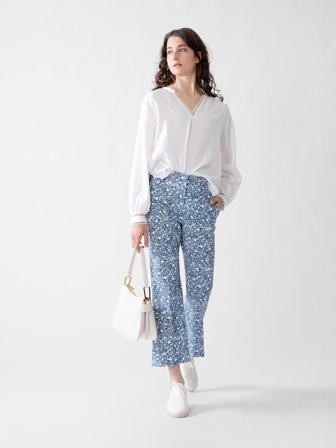 Eine Frau steht vor einem schlichten Hintergrund. Sie trägt eine weite weiße Bluse, eine blaue Hose mit Blumenmuster, weiße Turnschuhe und eine weiße Handtasche. Sie hat lockiges braunes Haar und blickt leicht zur Seite.