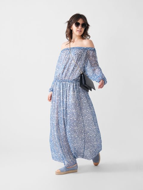 Eine Frau mit Sonnenbrille, einem blauen schulterfreien Maxikleid mit Blumenmuster, blauen Espadrilles und einer schwarzen Umhängetasche posiert selbstbewusst vor einem schlichten weißen Hintergrund.