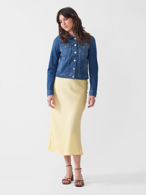 Eine Frau in einer blauen Jeansjacke, einem hellgelben Midirock aus Satin und Sandalen mit Leopardenmuster steht vor einem schlichten weißen Hintergrund. Sie blickt nach unten, die Hände hängen entspannt an den Seiten.