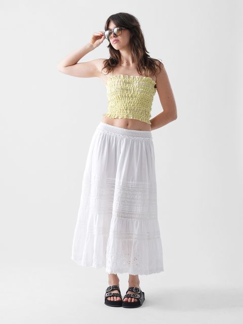 Eine Frau steht vor einem weißen Hintergrund. Sie trägt eine Sonnenbrille, ein gelbes trägerloses, gesmoktes Crop-Top, einen langen weißen Rock mit Spitzendetails und schwarze Pantoletten. Sie hat langes braunes Haar und posiert mit einer Hand auf ihrer Sonnenbrille.