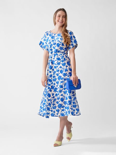 Vor einem schlichten Hintergrund steht eine lächelnde Frau. Sie trägt ein weißes Midikleid mit blauem Blumenmuster, hellgelbe offene Pumps und hält eine leuchtend blaue Clutch in der Hand.