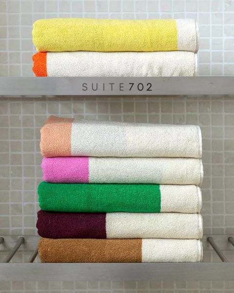Sechs ordentlich gefaltete Handtücher mit farbigen Enden – gelb, orange, braun, rosa, grün und weinrot – stapeln sich auf zwei Regalen vor einer gefliesten Wand. Auf dem oberen Regal steht der Schriftzug „SUITE702“.