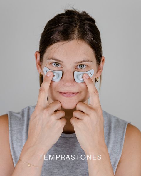 Eine Person hält zwei kleine, ovale Geräte unter den Augen und drückt sie sanft auf ihre Haut. Sie hat einen neutralen Gesichtsausdruck und trägt ein ärmelloses Oberteil. Am unteren Bildrand erscheint das Wort „TEMPRASTONES“.