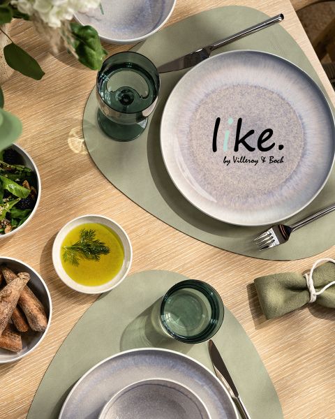 Draufsicht auf einen Esstisch mit einem weiß-blauen Teller mit der Aufschrift „like. by Villeroy & Boch“, grünen Tischsets, durchsichtigen Glasbechern, einer Schüssel mit Grissini, einer Schüssel mit Salat und einer Schüssel mit Öl, garniert mit Kräutern.