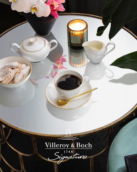 Auf einem Couchtisch aus reflektierendem Glas stehen eine Tasse Kaffee in einer weiß-goldenen Tasse, eine kleine Teekanne, ein Milchkännchen, eine Kerze und ein Teller mit Keksen. Die Szene wirkt elegant und gemütlich, und unten ist das Logo von Villeroy & Boch zu sehen.