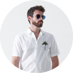 Ein Mann mit Bart, Sonnenbrille und einem weißen Kurzarmhemd mit einem kleinen Palmenmuster steht vor einem schlichten weißen Hintergrund.