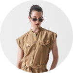 Eine Frau mit dunklem Haar zu einem Dutt trägt eine Sonnenbrille und ein hellbraunes, ärmelloses Kleid mit Knöpfen, Vordertaschen und Taillenband und steht vor einem schlichten weißen Hintergrund.