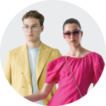Ein Mann in einem gelben Blazer und mit Brille steht neben einer Frau in einem leuchtend rosa Kleid und einer Sonnenbrille. Beide blicken nach vorne vor einem schlichten weißen Hintergrund.
