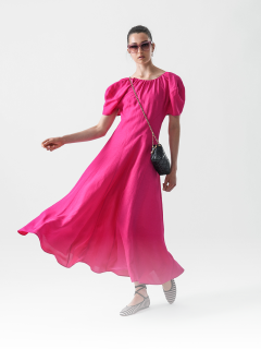 Eine Frau in einem leuchtend rosa, fließenden Kleid mit Puffärmeln posiert selbstbewusst. Sie trägt eine Sonnenbrille, eine kleine schwarze Umhängetasche und weiße, gestreifte flache Schuhe vor einem schlichten weißen Hintergrund.