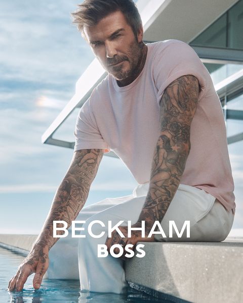 Ein tätowierter Mann in einem hellrosa T-Shirt und einer weißen Hose sitzt an einem modernen Pool und berührt das Wasser. Der Text „BECKHAM BOSS“ ist über das Bild gelegt.