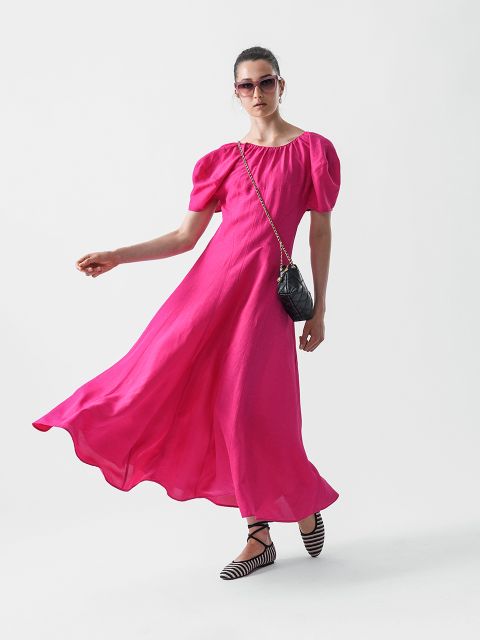 Eine Frau in einem leuchtend rosa, fließenden Kleid mit Puffärmeln, schwarz-weiß gestreiften Schuhen, einer schwarzen Umhängetasche und einer Sonnenbrille posiert vor einem schlichten weißen Hintergrund.