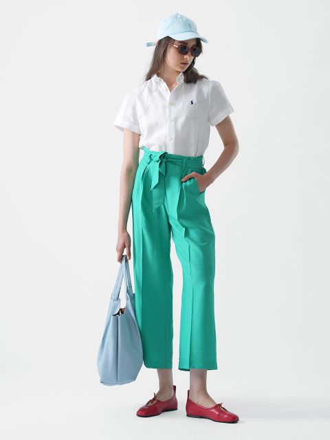 Eine Frau mit einer hellblauen Kappe, einer Sonnenbrille, einem weißen Kurzarmhemd, grünen Hosen mit hoher Taille und Gürtel, roten Slippern und einer hellblauen Einkaufstasche in der Hand steht vor einem schlichten weißen Hintergrund.