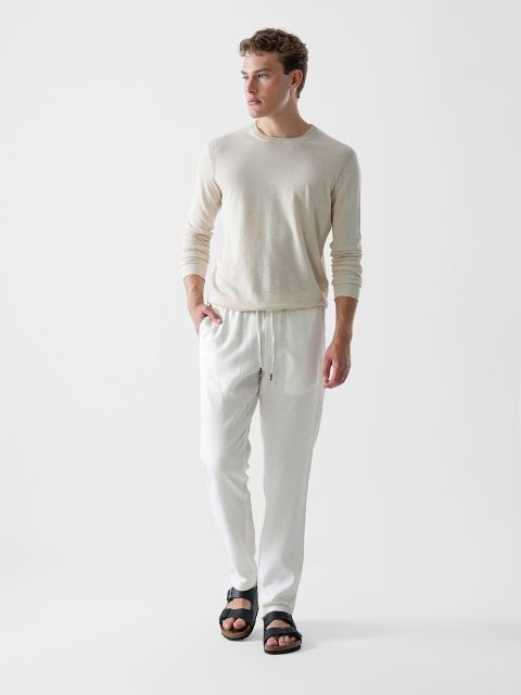 Ein Mann in einem hellbeigen Langarmhemd, einer weißen Hose mit Kordelzug und schwarzen Sandalen steht vor einem schlichten weißen Hintergrund und blickt leicht nach links.