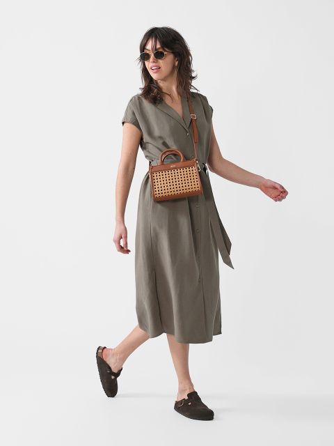 Eine Frau mit Sonnenbrille, einem kurzärmeligen olivgrünen Midikleid, braunen Slippern und einer kleinen braunen gewebten Umhängetasche läuft vor einem schlichten weißen Hintergrund.