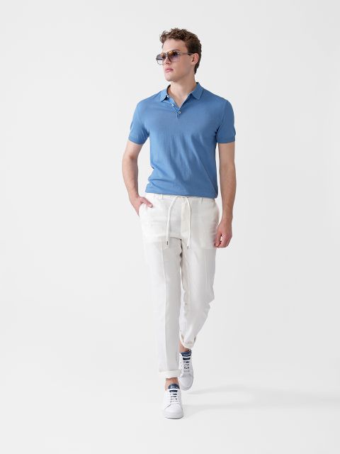 Ein Mann mit Sonnenbrille, hellblauem Kurzarm-Poloshirt, weißen Hosen mit Kordelzug und weißen Turnschuhen geht selbstbewusst vor einem schlichten weißen Hintergrund.