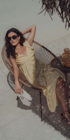 Eine Frau in einem hellgelben Kleid und mit Sonnenbrille sitzt im Freien in einem Korbstuhl und hält eine weiße Handtasche in der Hand. Sie sitzt entspannt, einen Arm hinter dem Kopf, umgeben von Sonnenlicht und Schatten auf dem Fliesenboden.