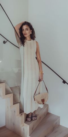Eine Frau in einem ärmellosen, langen weißen Strickkleid steht auf einer Holztreppe. Sie hält eine Strohhandtasche in der Hand und stützt einen Arm auf das Treppengeländer. Sie hat welliges dunkles Haar, trägt Sandalen und Goldschmuck.