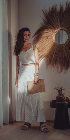 Eine Frau in einem passenden weißen Crop-Top und einem langen Rock steht drinnen und hält eine strukturierte beige Clutch in der Hand. Sie steht neben einer Wand mit einem dekorativen Sonnenspiegel und einem kleinen Tisch mit einer Vase und Zweigen. Sonnenlicht fällt herein.