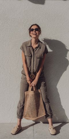 Eine Frau mit Sonnenbrille steht lächelnd vor einer hellen Wand. Sie trägt einen lockeren, olivgrünen Overall, beige Slipper und hält eine gewebte Einkaufstasche vor sich. Sonnenlicht wirft ihren Schatten an die Wand.