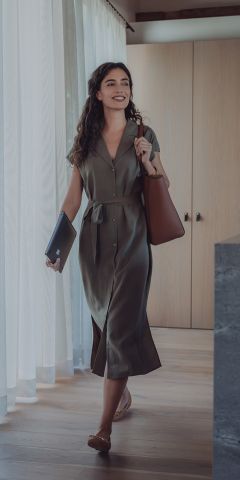 Eine Frau mit lockigem Haar, einem grünen Button-Down-Kleid und flachen Schuhen, lächelt, als sie ins Haus geht. Sie trägt eine braune Einkaufstasche und einen zugeklappten Laptop in der Hand. Licht fällt durch die transparenten Vorhänge neben ihr.
