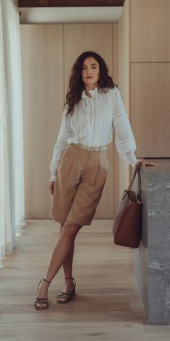 Eine Frau mit lockigem Haar, einer weißen Bluse, hellbraunen knielangen Shorts und Sandalen steht drinnen an einer Steintheke und hält eine braune Einkaufstasche in der Hand. Licht fällt durch transparente Vorhänge in den modernen, minimalistischen Raum.