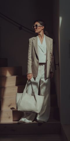 Eine Frau mit Sonnenbrille und beigem Blazer steht auf einer Holztreppe und hält eine große weiße Einkaufstasche in der Hand. Sie trägt eine weite weiße Hose und ein weißes Oberteil, das in dramatisches Licht und Schatten getaucht ist.