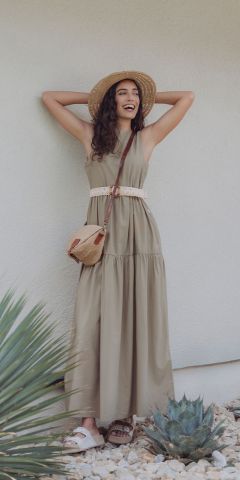 Eine Frau in einem ärmellosen olivgrünen Maxikleid, Strohhut und Umhängetasche steht lächelnd vor einer hellen Wand, zu ihren Füßen liegen Sukkulenten und Steine. Sie hat welliges Haar und trägt Sandalen.