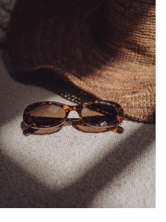 Eine Sonnenbrille aus Schildpatt liegt auf einer hellen Stoffoberfläche neben einem braun gewebten Sonnenhut, beide werden teilweise vom Sonnenlicht beleuchtet.
