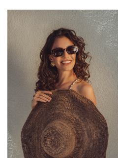 Eine lächelnde Frau mit lockigem Haar trägt eine Sonnenbrille und hält einen großen gewebten Sonnenhut vor einer neutral strukturierten Wand. Sie wirkt entspannt und stilvoll.
