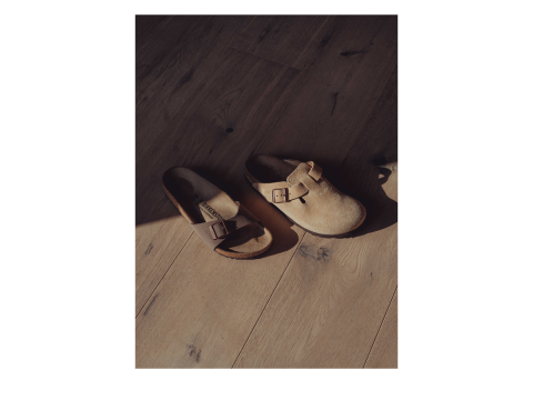 Zwei Slipper aus hellbraunem Wildleder mit Schnallen stehen auf einem Holzboden, teilweise im Schatten.
