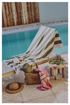 Ein gestreiftes Handtuch bedeckt eine Liege am Pool neben einem kleinen Tisch mit Getränken. In der Nähe stehen auf der Steinterrasse am Swimmingpool eine gewebte Tasche mit einem rosa Schal, ein Buch, eine Sonnenbrille und ein Strohhut.
