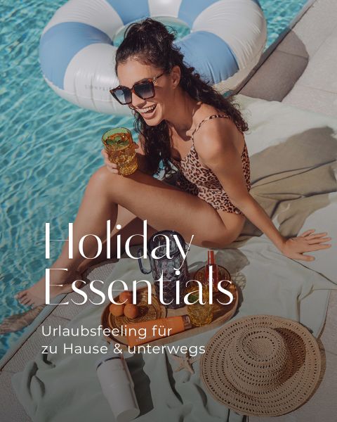 Eine Frau in einem Badeanzug mit Leopardenmuster sitzt lächelnd an einem Pool und hält einen Drink in der Hand. Sonnenbrillen, Sonnenhut, Sonnencreme und Obst sind in der Nähe. Text lautet: „Holiday Essentials. Urlaubsfeeling für zu Hause & unterwegs.“.