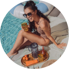 Eine Frau in einem Badeanzug mit Leopardenmuster sitzt lächelnd auf einem Handtuch am Pool und hält ein Getränk in der Hand. Sie trägt eine Sonnenbrille und steht neben einem Tablett mit Getränken, Sonnencreme und Obst. Hinter ihr ist im Wasser eine aufblasbare Schwimmhilfe zu sehen.