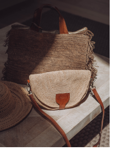 Zwei geflochtene Handtaschen, eine große und eine kleine mit Schultergurt, beide mit braunen Lederakzenten, liegen auf einer Holzfläche neben einem teilweise sichtbaren geflochtenen Hut. Sonnenlicht wirft weiche Schatten auf die Szene.