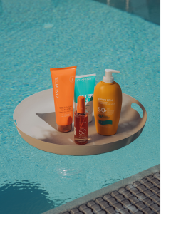 In einem Pool schwimmt ein rundes beiges Tablett, auf dem sich vier Sonnenschutzprodukte in orange- und türkisfarbenen Verpackungen befinden. In der unteren rechten Ecke ist ein Poolrand zu sehen.