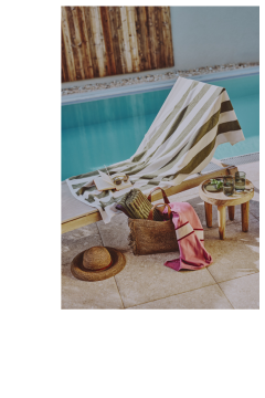 Eine Szene am Pool mit einem Liegestuhl, der mit einem gestreiften Handtuch bedeckt ist, einem Sonnenhut auf dem Boden, einer Strohtasche mit einem rosa Schal, einer Sonnenbrille und einem kleinen Tisch mit Getränken und einem Teller Obst.
