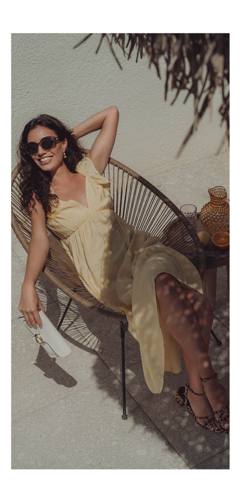 Eine Frau in gelbem Kleid und Sonnenbrille entspannt sich in einem geflochtenen Stuhl im Freien und lächelt in die Sonne. Sie hält eine weiße Clutch in der Hand, neben ihr auf einem kleinen Tisch ein Getränk und ein Krug.