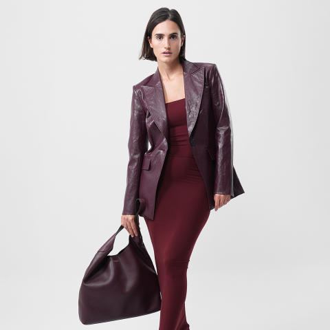 Eine Frau in einem taillierten weinroten Kleid und einem passenden Lederblazer trägt eine große weinrote Handtasche. Sie steht selbstbewusst vor einem schlichten hellen Hintergrund und blickt direkt in die Kamera.