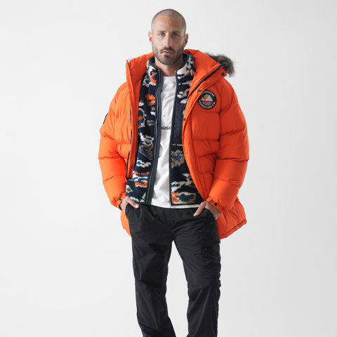 Ein Mann steht vor einem schlichten weißen Hintergrund und trägt eine leuchtend orangefarbene Pufferjacke mit einer pelzbesetzten Kapuze, einen gemusterten Schal, ein weißes Hemd und eine schwarze Hose. Er schaut mit neutralem Blick direkt in die Kamera.