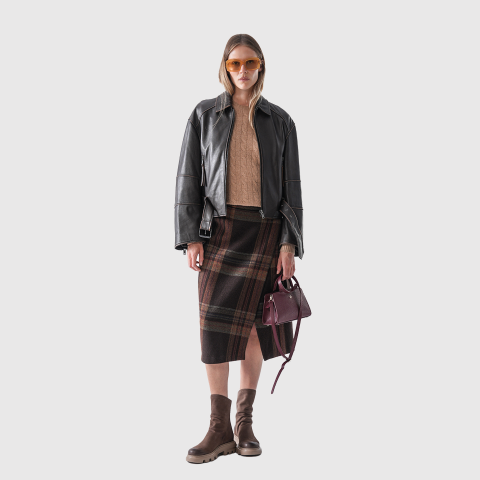 Eine Frau mit Sonnenbrille, schwarzer Lederjacke, beigem Pullover, kariertem Rock und braunen Stiefeln hält eine kleine weinrote Handtasche und steht vor einem schlichten hellen Hintergrund.