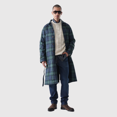Ein Mann mit Sonnenbrille, einem langen grün-blau karierten Mantel über einem cremefarbenen Pullover und blauen Jeans, mit braunen Stiefeln, steht vor einem schlichten weißen Hintergrund.