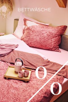 Ein ordentlich gemachtes Bett mit rosa geblümtem Bettzeug, einer gemusterten roten Decke und einem Holztablett mit einer Teekanne und zwei Tassen. Das Wort "BETTWÄSCHE" und große Prozentzeichen überlagern das Bild.