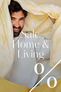 Ein lächelnder Mann in einem weißen Hemd sitzt unter einer gelb gemusterten Decke. Der überlagerte weiße Text lautet: "Sale Home & Living" mit einem großen Prozentzeichen, das auf einen Haushaltswarenverkauf hindeutet.