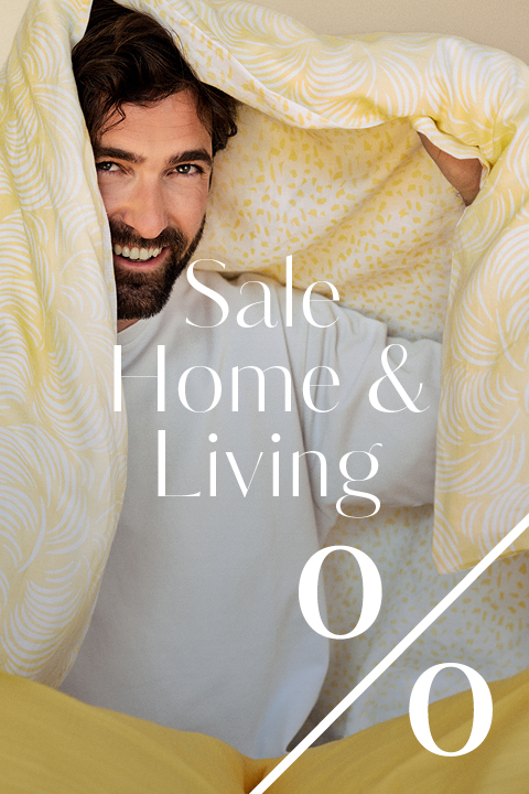 Ein lächelnder Mann in einem weißen Hemd sitzt unter einer gelb gemusterten Decke. Der überlagerte weiße Text lautet: "Sale Home & Living" mit einem großen Prozentzeichen, das auf einen Haushaltswarenverkauf hindeutet.