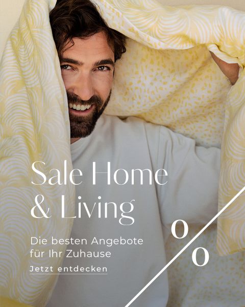 Ein lächelnder Mann mit Bart hält eine gelb gemusterte Decke hoch, in die er sich teilweise einwickelt. Der Text lautet: "Sale Home & Living. Die besten Angebote für Ihr Zuhause. Jetzt entdecken.